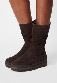 Braune Wildleder-Stiefeletten mit gerafftem Design, flacher Plateausohle und seitlichem Reißverschluss. Glatte Textur und minimale Nähdetails.