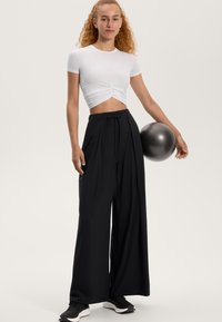 Valkoinen lyhyt toppi, jossa on rypytysdetaljeita, yhdistettynä löysiin mustiin housuihin, jotka on kiristetty vyötäröltä. Malli pitää mustaa kuntopalloa.