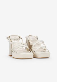 Sandalias de plataforma de charol blanco con tacón grueso, múltiples correas entrecruzadas y un hebilla en el tobillo trasero. Textura suave con un acabado brillante.