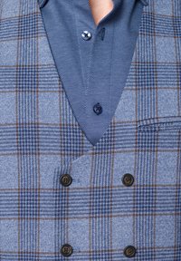 Niebieska kamizelka w kratkę o teksturowanej powierzchni, z brązowymi akcentami, podwójnymi guzikami oraz kontrastową niebieską koszulą z wzorem pod spodem.