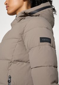 Lichtbeige pufferjack met een gestructureerd oppervlak, afgewerkt met horizontaal gestikte secties en een zwart logo op de linkerarm.