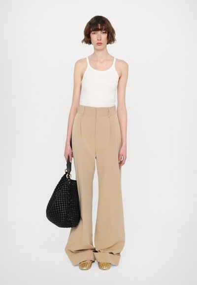 Beige weit geschnittene Hosen mit hoher Taille, kombiniert mit einem weißen Tanktop. Eine schwarze gewebte Schultertasche und goldene Slipper runden das Outfit ab.