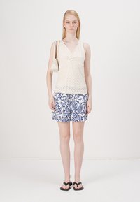 Topp med blondermønster i kremfarge, marineblå og hvite blomstermønstrede shorts, svarte flip-flops og en liten hvit håndveske.