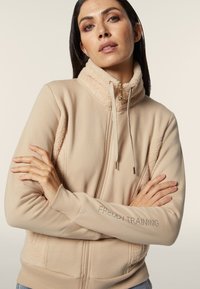 Giacca beige con zip, caratterizzata da un colletto in felpa e pannelli laterali, chiusura con cordino e ricamo "FREDDY TRAINING" sulla manica.