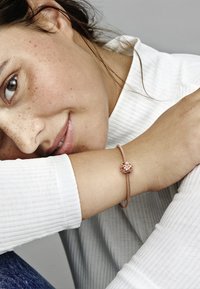 Bracciale in oro rosa con una fascia testurizzata e un charm floreale con accenti rosa e trasparenti. Indossato su un polso, abbinato a un top bianco a coste.
