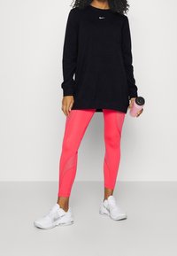 T-shirt noir à manches longues Nike, leggings roses avec accents en mesh, et baskets blanches avec des détails gris. Tenant une bouteille d'eau rose.