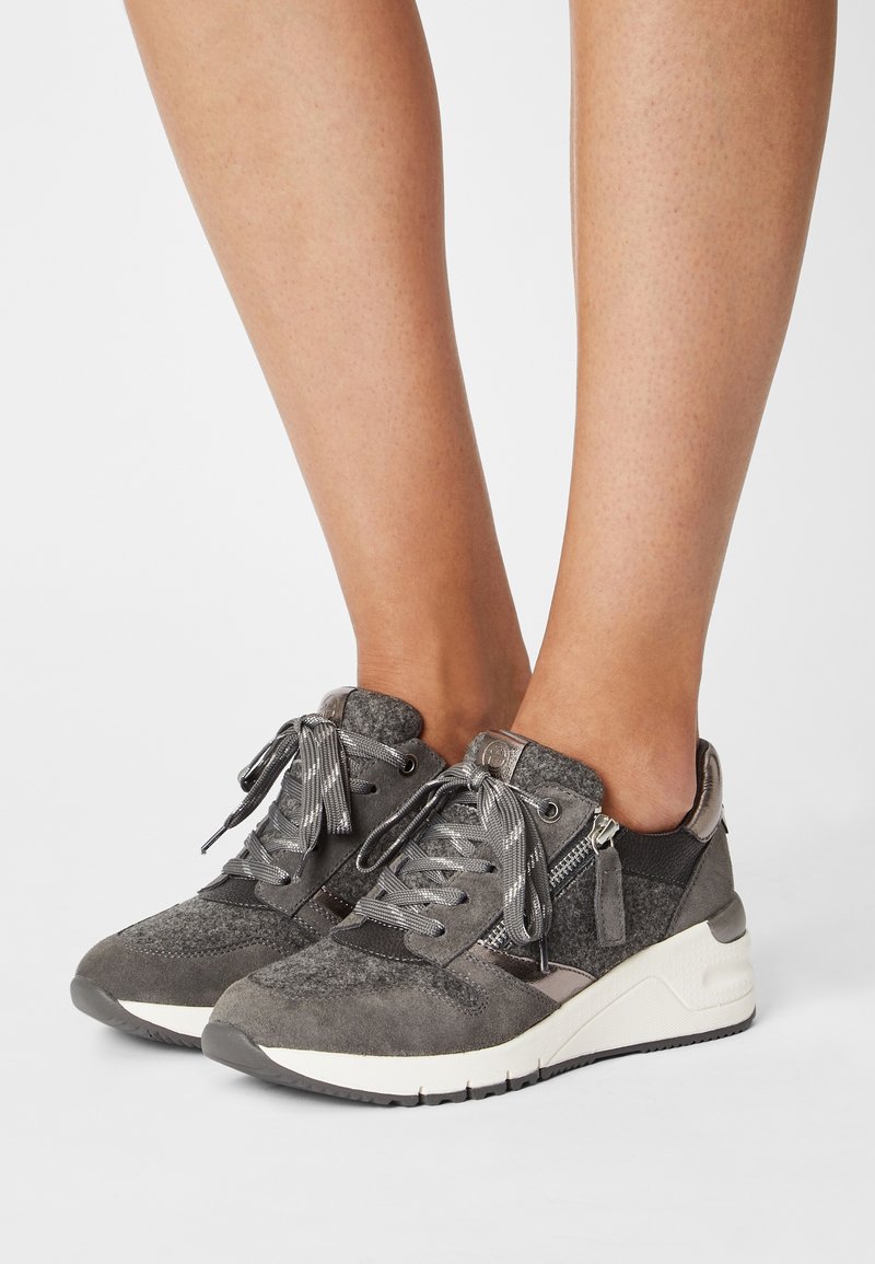 Pieds portant des baskets en daim gris et tissu avec semelles blanches épaisses, lacets et détails de fermeture éclair latérale sur fond blanc.