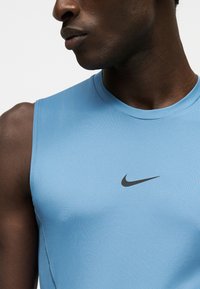 Homme portant un débardeur sportif Nike bleu clair, visible de l'épaule au cou, sur un fond uni.