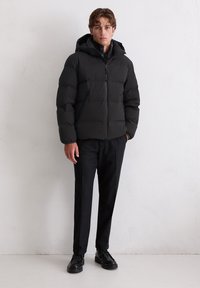 Schwarze Puffjacke mit Kapuze und Frontreißverschluss, kombiniert mit dunkel strukturierten Hosen und schwarzen Schuhen, vor einem neutralen Hintergrund.