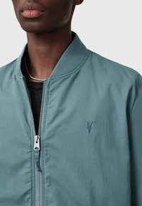 Giacca bomber in teal chiaro realizzata in tessuto morbido, con zip frontale e un logo ricamato discreto sul petto. Texture liscia, design casual.