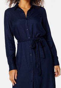 Vestito camicia blu navy con bottoni, maniche lunghe, vita aderente, texture liscia e leggero lucido, con bottoni abbinati.