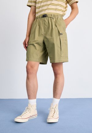 Persona que lleva pantalones cortos cargo verde oliva con bolsillos laterales, zapatillas de deporte altas beige, calcetines blancos y una camiseta de rayas de manga corta.