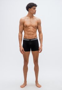 Νεαρός μυώδης άνδρας στέκεται ξυπόλυτος, φορώντας μαύρα εσώρουχα Jack & Jones, κοιτάζει προς τα δεξιά του πάνω σε απλό φωτεινό φόντο.