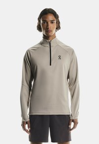 Šviesiai rudų spalvų sportinė marškinė ilgomis rankovėmis su užtrauktuku ant apykaklės, su juodu logotipu kairėje krūtinės pusėje ir tekstūruotomis rankovėmis. Dėvima su tamsiomis šortais.