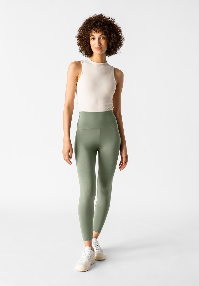Top in maglia a coste beige chiaro abbinato a leggings verdi oliva a vita alta, morbidi, completato da sneakers bianche, su uno sfondo semplice.
