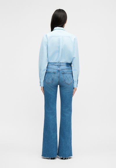 Žena s dlhými tmavými vlasmi nosiaca svetlomodrú košeľu zastrčenú do vysokých, rozšírených modrých džínsov s ošúchanými lemy, stojaca otočená chrbtom.