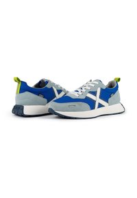 Zapatillas azules y grises con parte superior de ante y malla, detalles en blanco, suela de goma y un tirador verde en el talón.