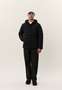 Mann som står iført en svart boblejakke, mørke bukser, svarte sko og en hvit baseballcap foran en ensfarget beige bakgrunn.