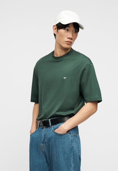 Άνδρας που φοράει σκούρο πράσινο κοντομάνικο μπλουζάκι, ανοιχτό μπλε τζιν με μαύρη ζώνη και λευκό καπέλο μπέιζμπολ, με τα χέρια στις τσέπες, κοιτάζοντας προς τα πλάγια.