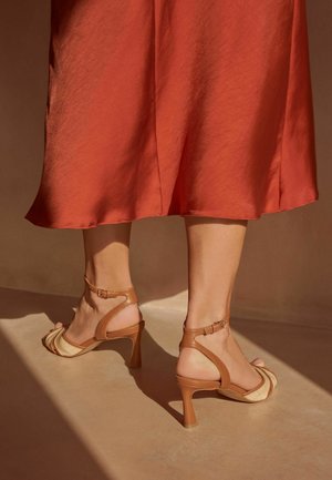 Beine mit hellbraunen Sandalen mit Absatz und Knöchelriemen sowie einem rostfarbenen Midirock, stehend auf einem sonnenbeschienenen Boden.