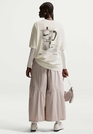 Persona con cabello trenzado que lleva una camiseta blanca de gran tamaño con un gráfico de cafetera y taza, pantalones anchos beige y sostiene una bolsa beige.