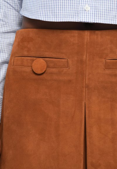 Gonna in suede marrone con due tasche orizzontali, ciascuna dotata di un bottone tondo. Il tessuto ha una texture morbida, decorata con linee sartoriali e uno spacco.