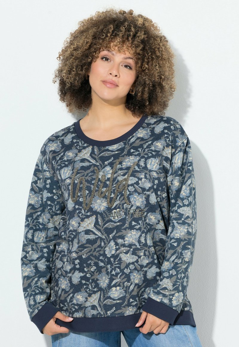 Mujer con cabello rizado vistiendo un sudadera floral azul marino con la palabra "Wild" escrita en ella, posando frente a un fondo blanco liso.