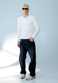 Blonde Person trägt schwarze Sonnenbrille, weißes Langarmshirt, lockere dunkle Jeans und weiße Sneaker und steht vor einem schlichten hellen Hintergrund.