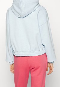 Ljusblå kort hoodie med dragsko i huvan och elastiskt midjeband, kombinerad med rosa sweatpants. Tyget verkar mjukt och strukturerat.