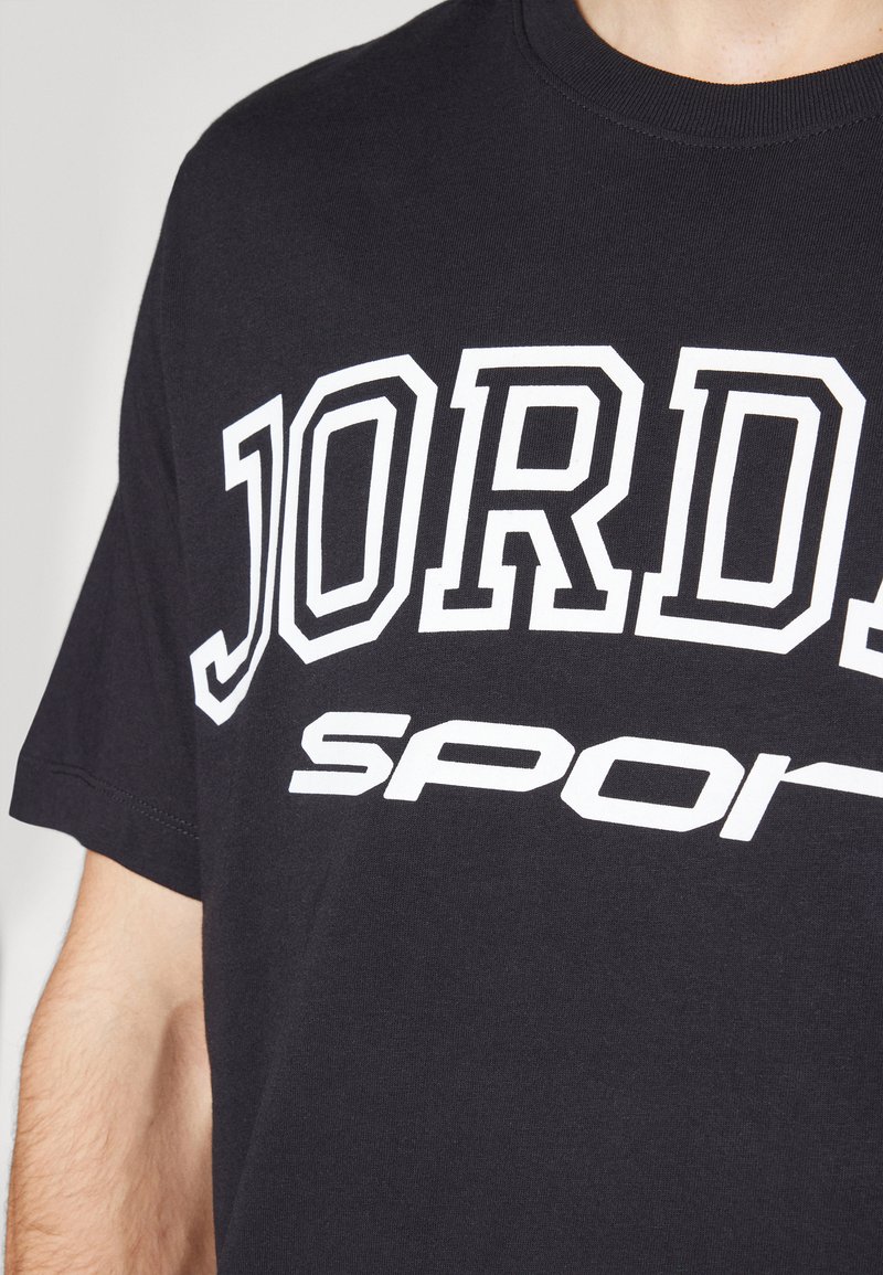 Camisa negra de manga corta con un gran texto blanco "JORD" y un texto "sport" parcialmente visible, llevada por una persona mostrada de cuello a mitad del torso.