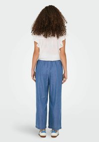 Top blanco con lunares negros y mangas acampanadas, combinado con jeans de pierna ancha azules. La modelo es vista desde atrás, usando zapatillas blancas con detalles azules.