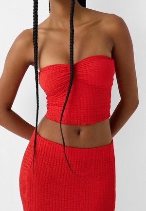 Vrouw met lange vlechten, gekleed in een felrode textuur strapless crop top en bijpassende hoge taille rok, staande tegen een effen achtergrond.