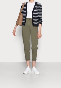 Randig marinblå kofta med guldfärgade knappar över en vit t-shirt, olivgröna korta byxor och vita sneakers, kombinerat med en brun handväska.