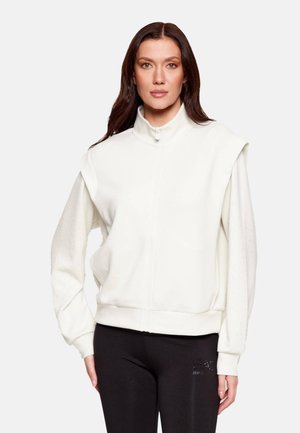 Donna con lunghi capelli scuri che indossa una felpa bianca con cerniera e spalle imbottite e pantaloni neri aderenti su uno sfondo semplice.