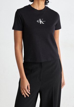 Person wearing a fitted black short-sleeve Calvin Klein Jeans t-shirt and loose black pants against a plain light background.