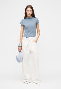 Femme portant un haut bleu clair et un pantalon large blanc, tenant un sac bleu assorti et un accessoire de poignet, debout devant un fond uni.