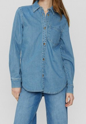 Frau trägt ein hellblaues Denimhemd mit Knöpfen und Brusttasche, kombiniert mit passenden blauen Denim-Jeans.