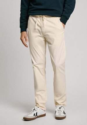 Pantalones beige con cintura elástica y cordón, que cuentan con bolsillos laterales y una silueta ajustada, combinados con zapatillas blancas.