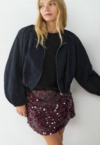 Schwarze, taillierte Bomberjacke mit voluminösen Ärmeln, kombiniert mit einem figurbetonten, burdeos-farbenen Minirock aus Pailletten, der eine glänzende, strukturierte Oberfläche aufweist.