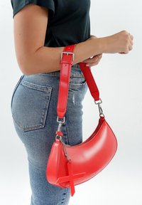 Borsa a mano in pelle rossa lucida con forma curva, tracolla rimovibile e un accento decorativo a forma di croce. Presenta una chiusura con cerniera e hardware in metallo.
