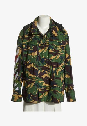 Veste style militaire avec motif camouflage vert, marron et beige, poches avant et broderie florale sur la manche gauche.