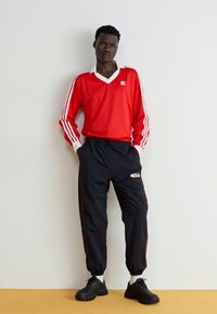 Rød langærmet skjorte med hvide striber og krave; sorte vindjakke bukser med elastiske manchetter; sorte sneakers.