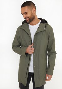 Veste imperméable vert olive avec capuche, fermetures à pression et poches latérales, portée sur un t-shirt blanc ; texture lisse et design moderne.