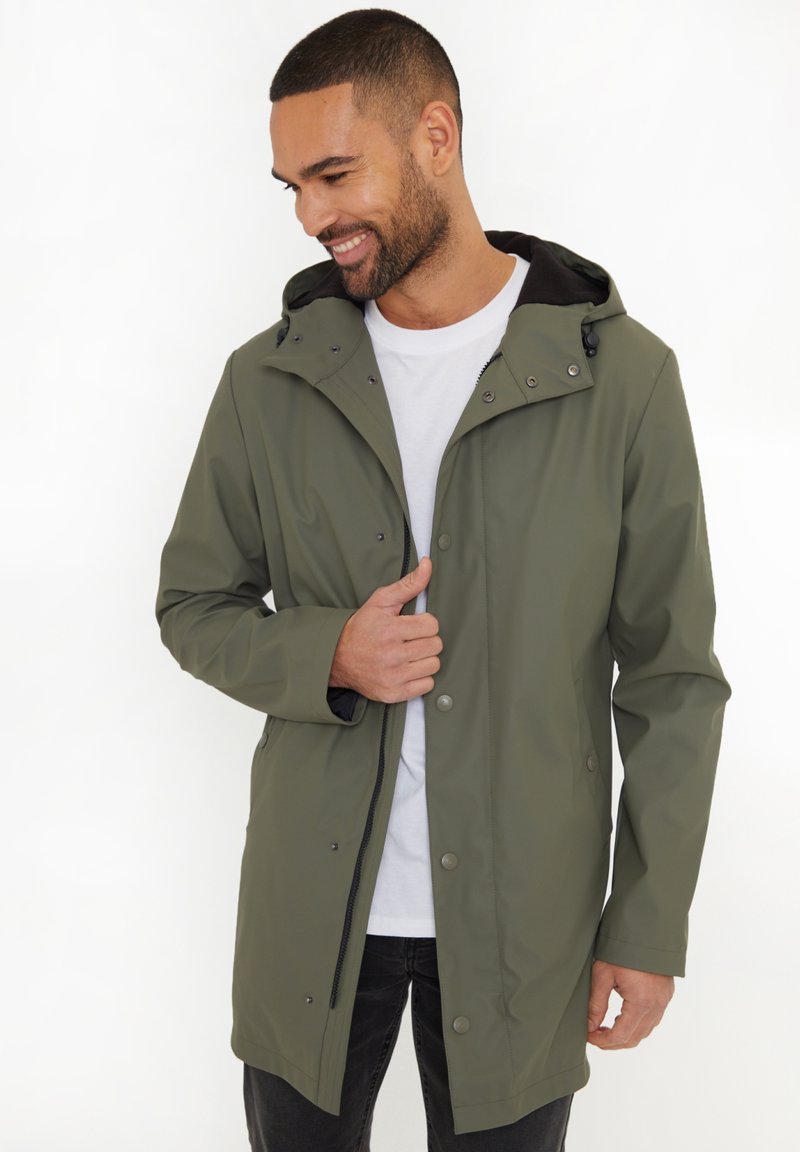 Veste imperméable vert olive avec capuche, fermetures à pression et poches latérales, portée sur un t-shirt blanc ; texture lisse et design moderne.