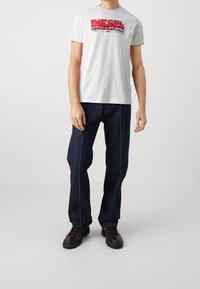 Hombre con camiseta blanca de la marca Diesel, jeans azul oscuro con costuras frontales y zapatos de cordones marrón oscuro, de pie frente a un fondo liso.