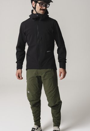 Man wearing black helmet, black jacket, green pants, and white sneakers standing against a plain light background.
