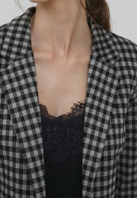 Kobieta w czarnej koszulce na ramiączkach z koronkowym wykończeniem pod szarym i czarnym w kratę żakietem, eksponująca dekolt i obojczyki.