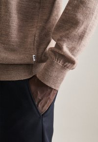 Brauner Strickpullover mit gerippten Bündchen, der eine strukturierte Oberfläche zeigt. Kombiniert mit dunklen Hosen, bei denen eine Tasche teilweise sichtbar ist.