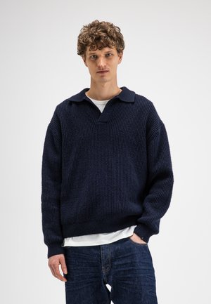 Young man with curly hair wearing a dark blue knit sweater over a white shirt and blue jeans, standing against a plain light background.