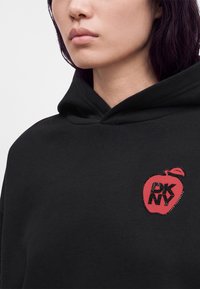 Gros plan d'une personne portant un sweat à capuche noir avec un logo pomme rouge comportant le texte « "DKNY" » sur la poitrine.